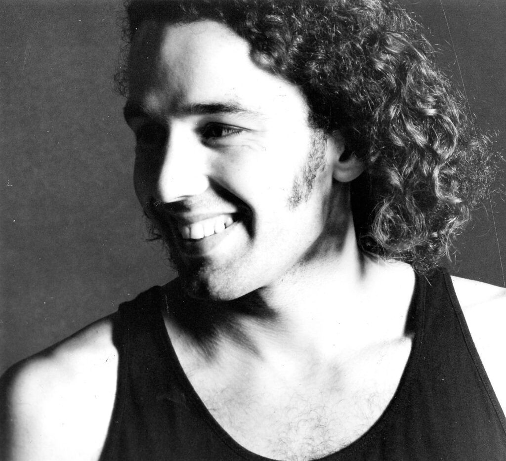Black and white photo of a 30 something male with curly, shoulder length hair and a big toothy smile is looking offscreen to picture left. The right side of his face is in shadow. He is wearing a black sleeveless undershirt.